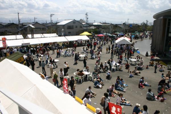 青空の下にたくさんのテントが並び、テントの外で地面に座っている人たちの写真
