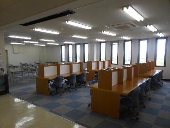 グレーと青色の市松模様のカーペットが敷かれ、中央に間仕切りがされ片面に3人ずつ座れる机が4台設置されている4階学習室の写真