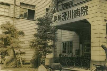 入口手前に数本の木々が植えられ、建物入口上部には「農協滑川病院」と書かれている農協滑川病院の外観を写したセピア色をした写真