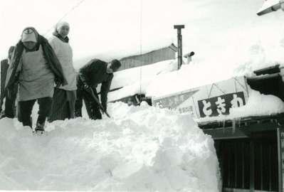 3名ほどの人が屋根部分まで積もった雪の上で雪かきをしている様子の白黒写真