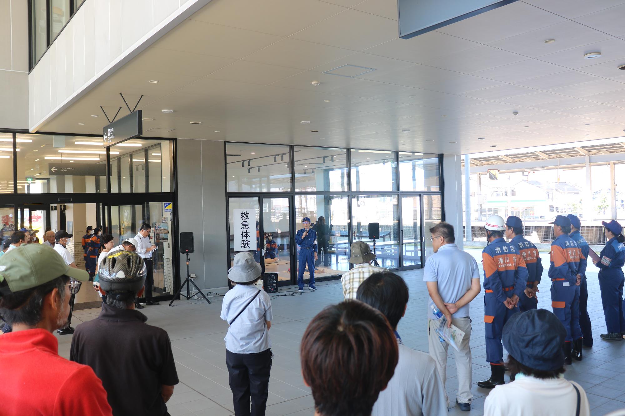 活動服を着た関係者の男性がマイクを持って話をしており、参加者の方々や関係者の方々が集まり話を聞いている様子の写真