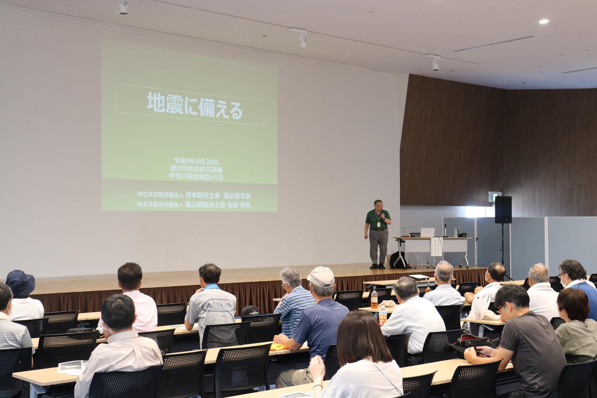 会場の前方にあるスクリーンに資料が写し出され、舞台横で説明する男性と多くの参加者の方々が席に着き、話を聞いている様子の写真