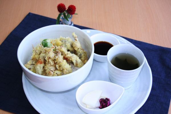 青色のランチョンマットが敷かれた白いお皿の上に、ほたるいか天丼とソース、スープ、漬物の皿がそれぞれ乗っている写真