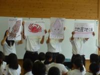 生徒が4人前に出て地産地消や海産物が描かれた紙を顔の前に持っている写真