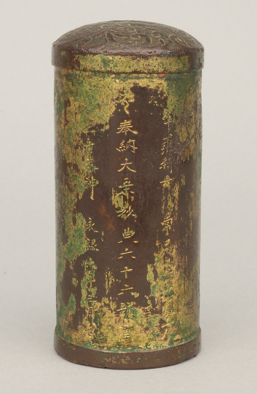 円筒状で蓋の上部は丸みを帯びており、筒の胴体には文字が刻まれ、模様が施されている銅製経筒の写真