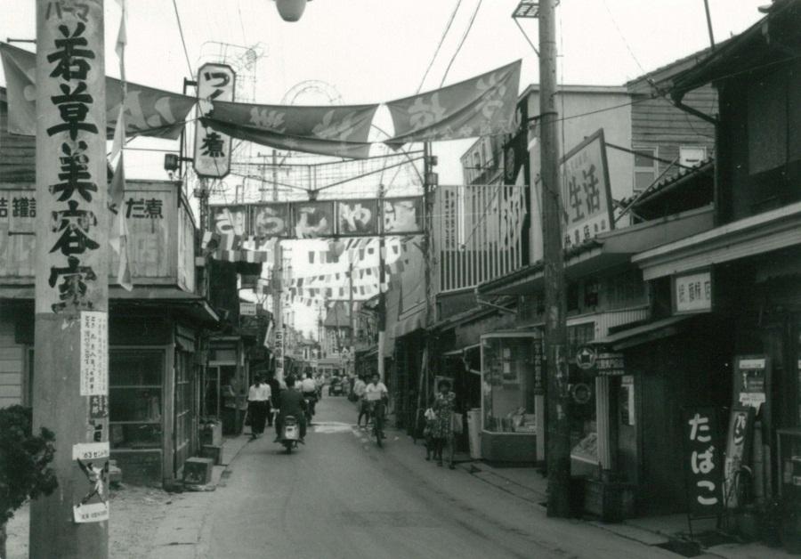 道路の両側に商店街が並んでおり、人々が行き来している通りの上に「さらしや通り」と書かれた看板が通路上に架かっている様子を写した白黒写真